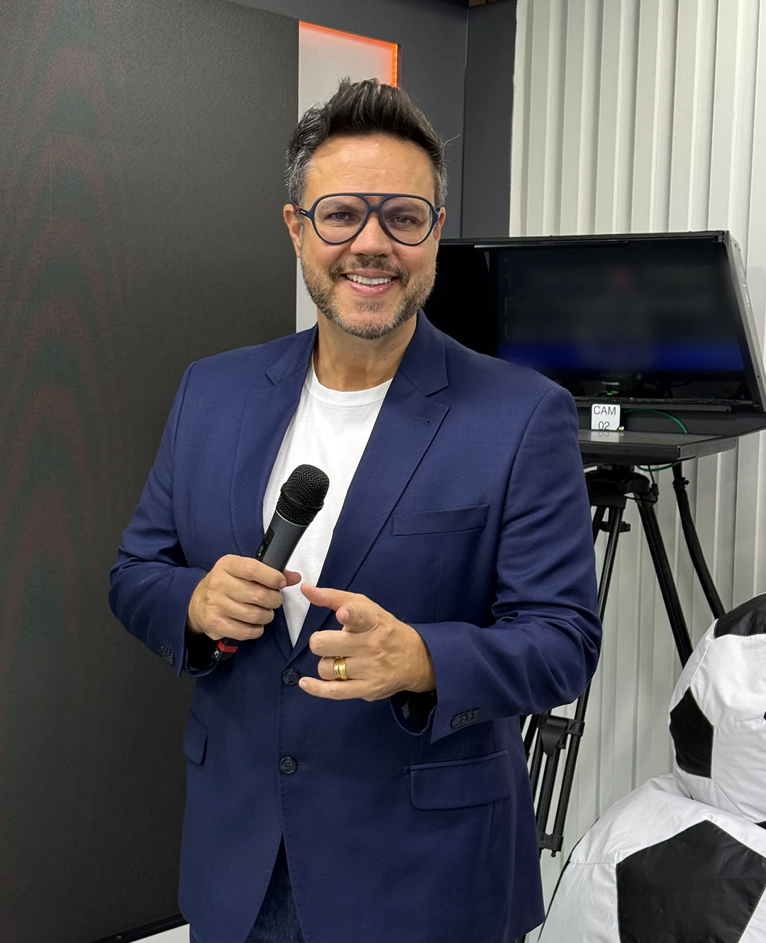 Vinícius Valverde de blazer, com microfone na mão, em um estúdio de TV